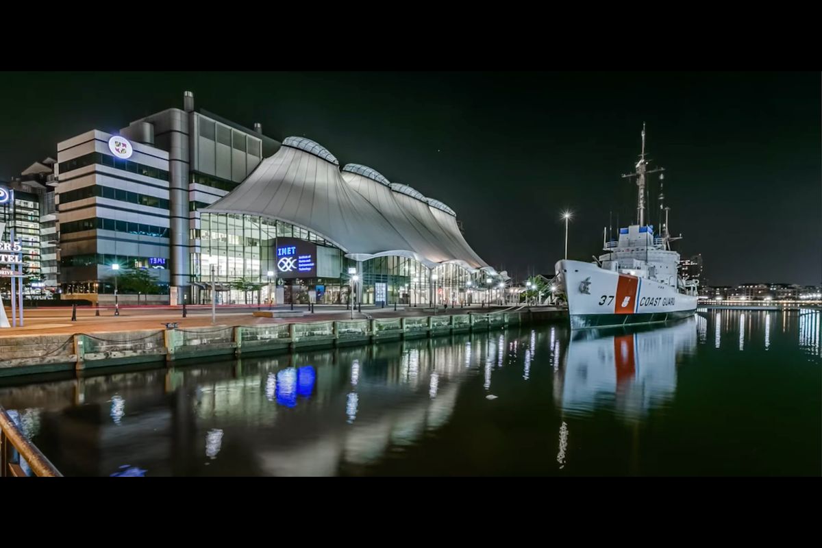 A nighttime view of a modern waterfront building with a curved roof next to a docked naval ship, illuminated by surrounding lights.