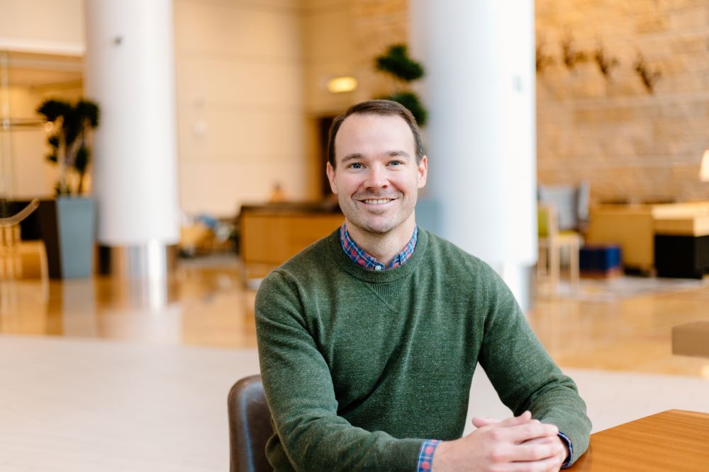 Man smiling at the camera while seated in a lobby with a relaxed posture.
