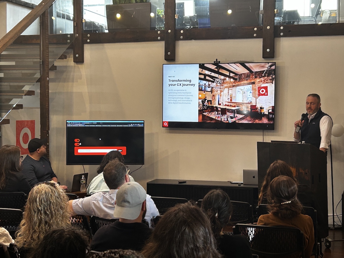 A presenter speaks at a conference in front of an audience with a presentation screen displaying company information behind him.