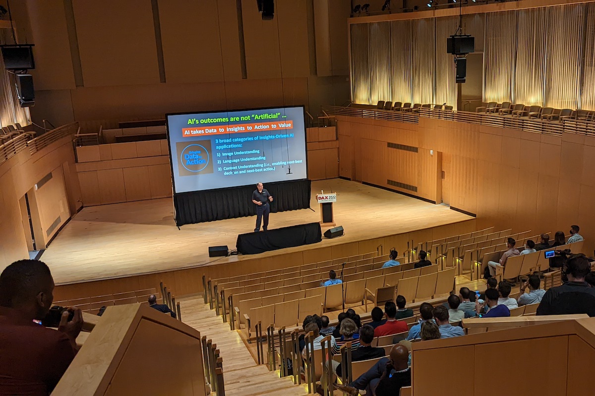 A speaker presenting at a conference in an auditorium with a slide titled "ai's outcomes are not 'artificial'" displayed on the screen.
