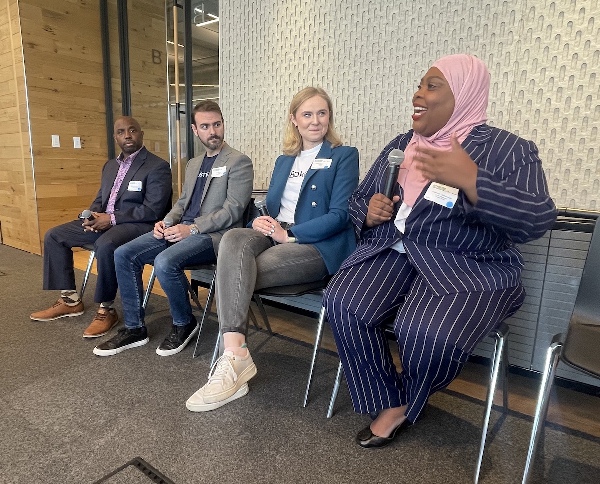 Four professionals sitting in a panel discussion, with one panelist speaking and the others listening.