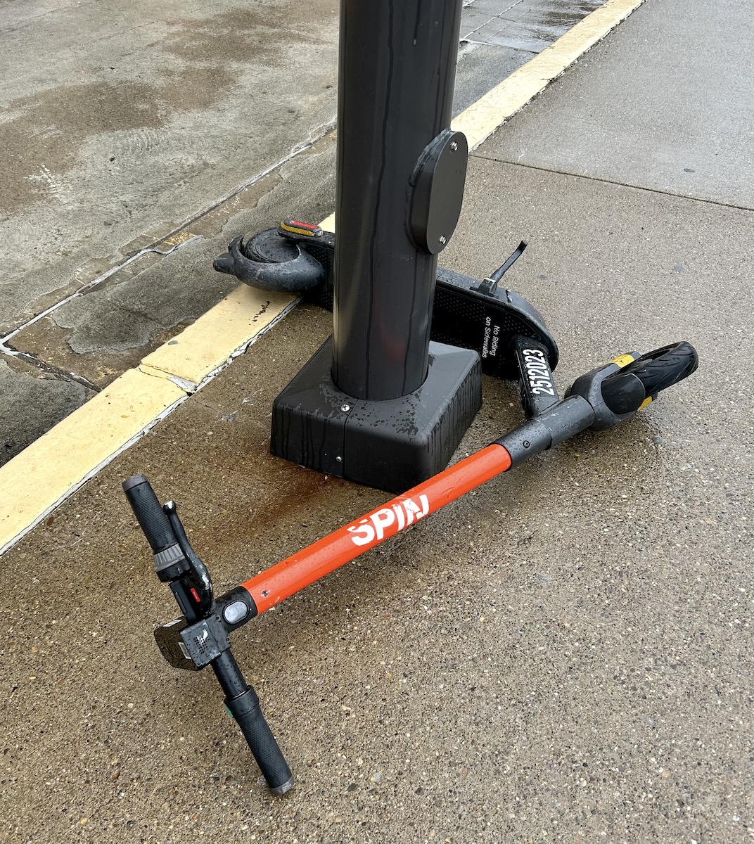 An electric scooter labeled "spin" lying on a wet pavement next to a pole.