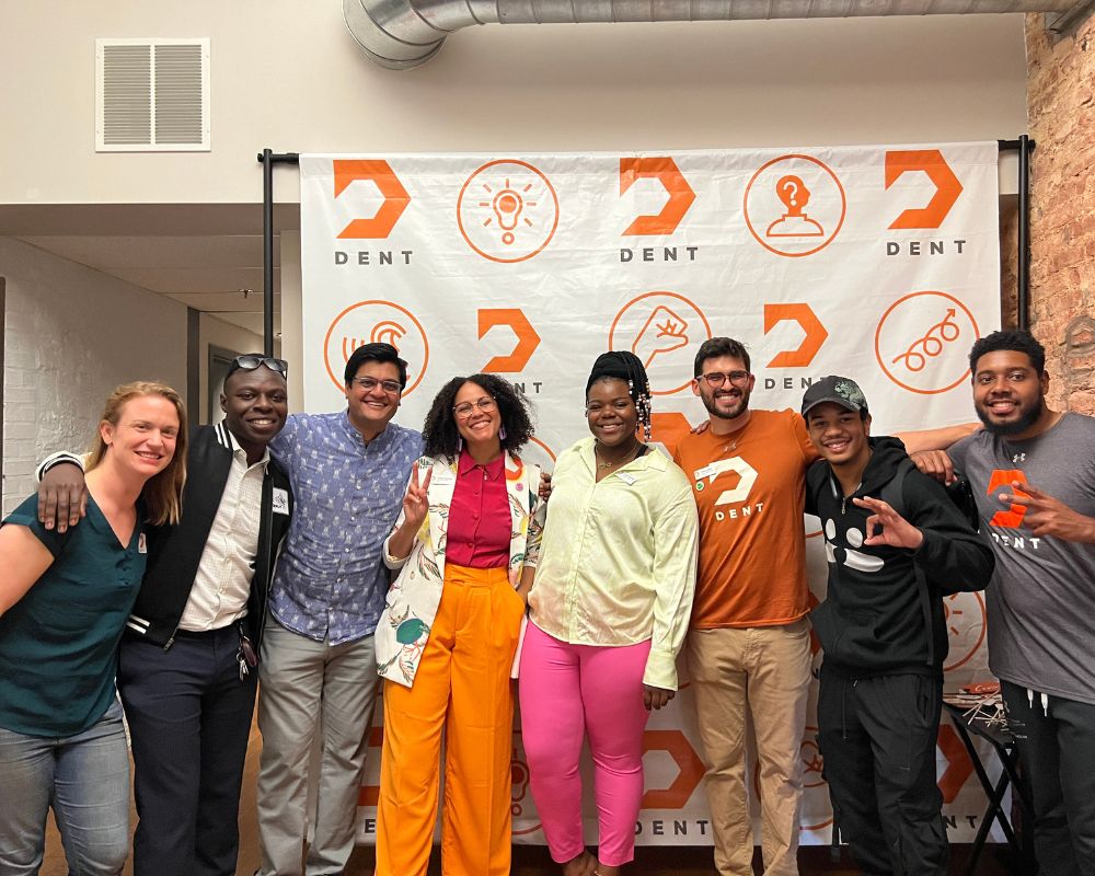 A group of eight individuals posing for a photo at an event with a backdrop that features the 'dent' logo.