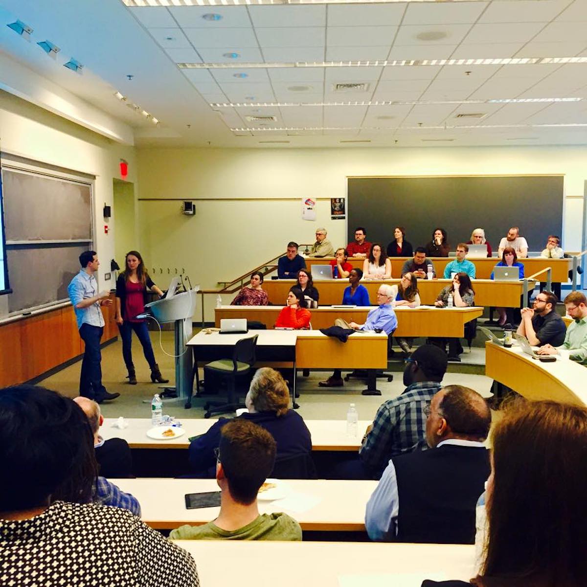 Two presenters speaking in front of a lecture hall filled with attending students and professionals.