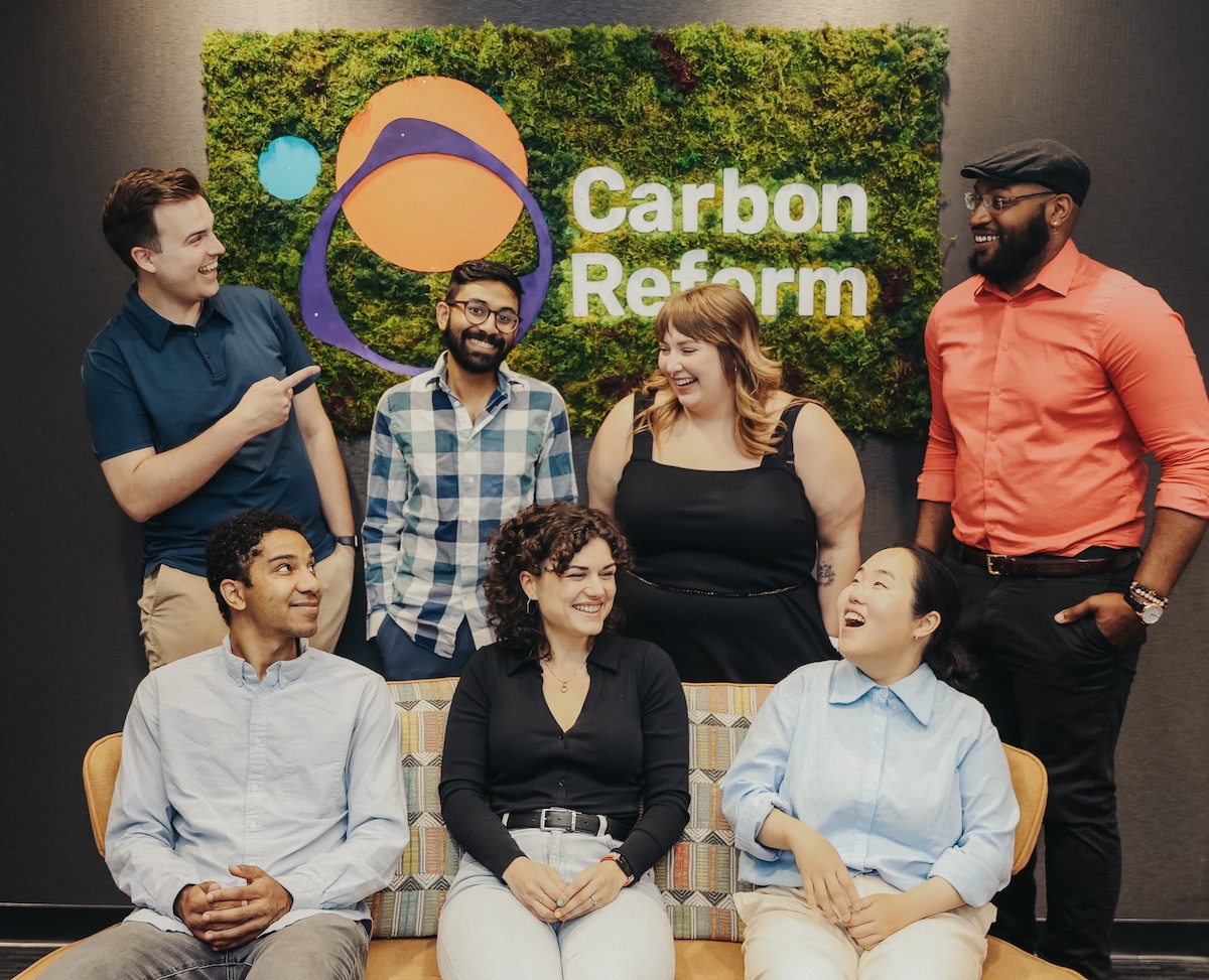 Seven people sit or stand in a huddle and smile in front a sign that says "Carbon Reform."