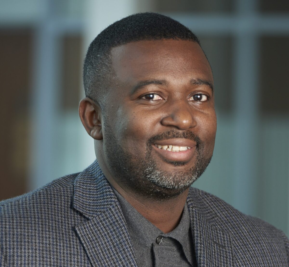 A smiling man wearing a blazer over a dark shirt, with a blurred background.
