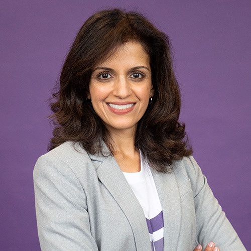 A woman in a grey blazer with her arms crossed in front of a purple background.