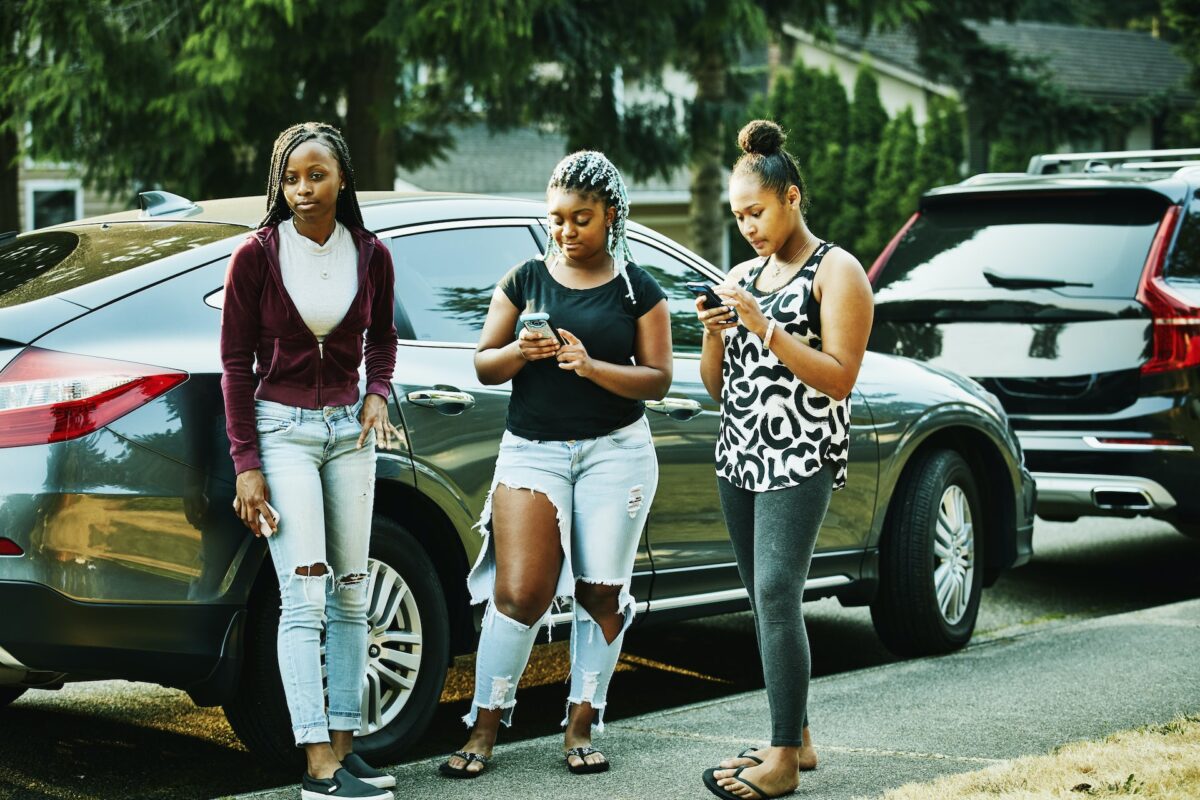 Three young women standing in front of a parked car looking at their phones.