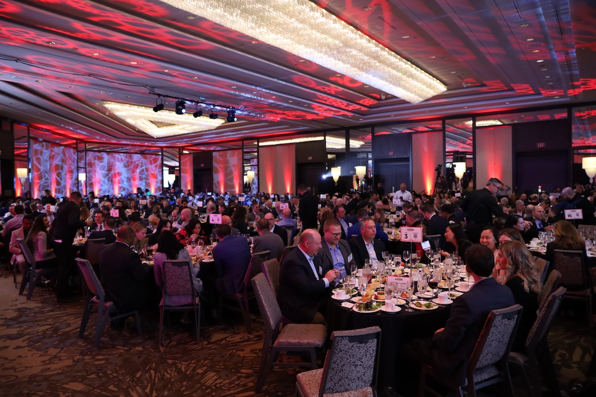 A crowd of people sitting at round tables with red and purple lighting.