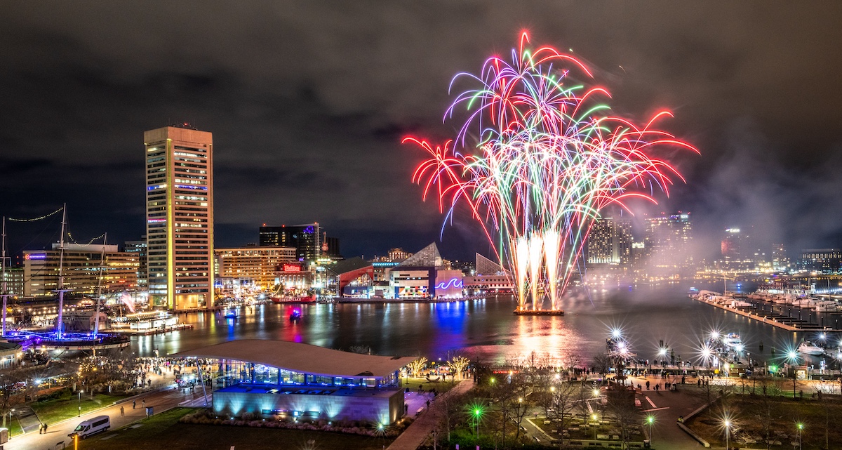 Fireworks light up the night sky over a city waterfront, with buildings and a marina in view.