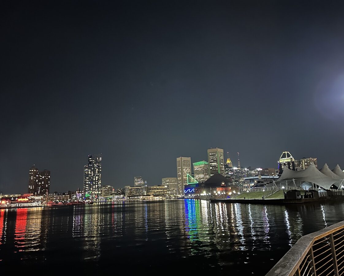 Nighttime view of a city skyline with illuminated buildings reflected on the water, including a white tent-like structure on the right, and vibrant colored lights along the waterfront.
