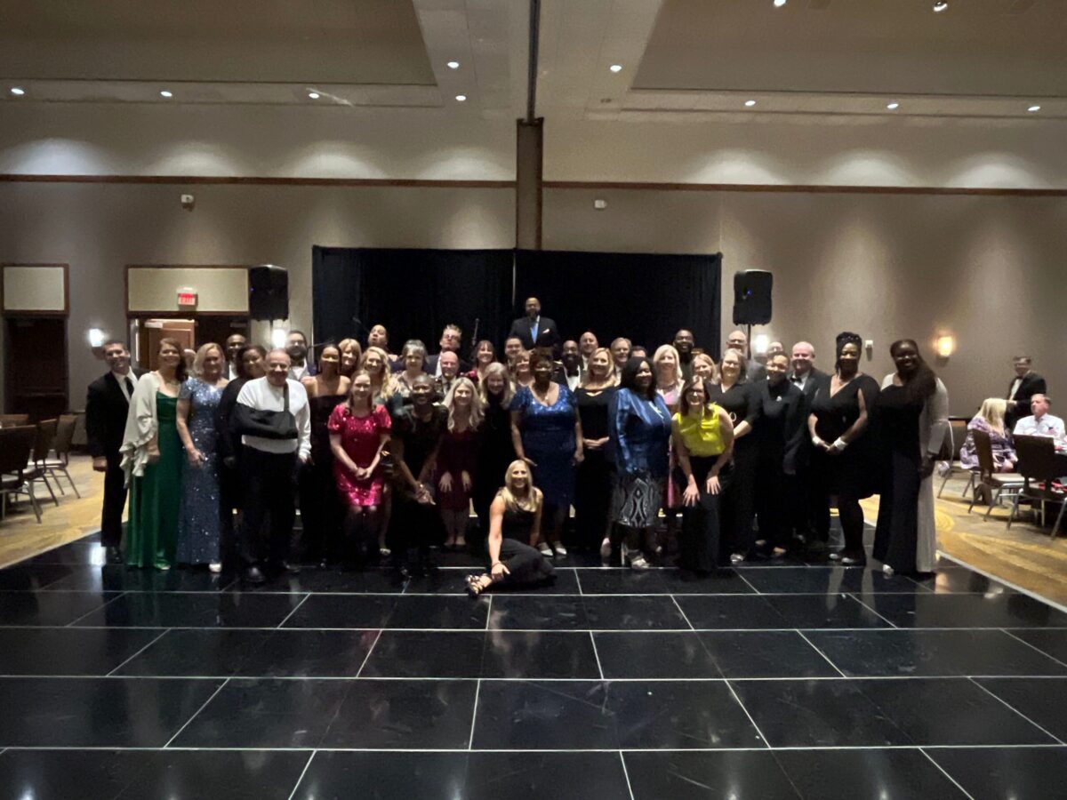 A large group of people dressed in formal attire pose together on a black-tiled dance floor in a well-lit indoor venue. One person is kneeling at the front.