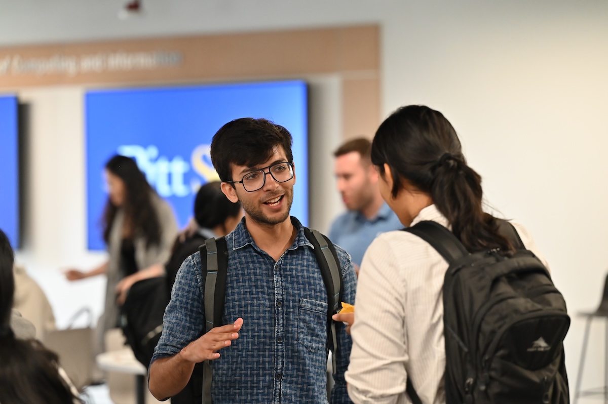 A man and woman with backpacks engage in conversation in a bright room, with a blurred background showing other people and a screen displaying the text "Pitt.