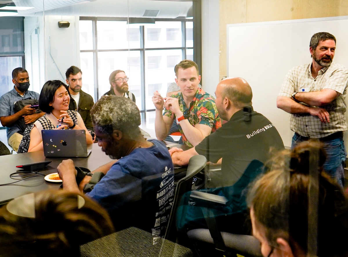 A diverse group of people in a meeting room, some are seated around a table with laptops and notebooks, while others stand and engage in conversation.