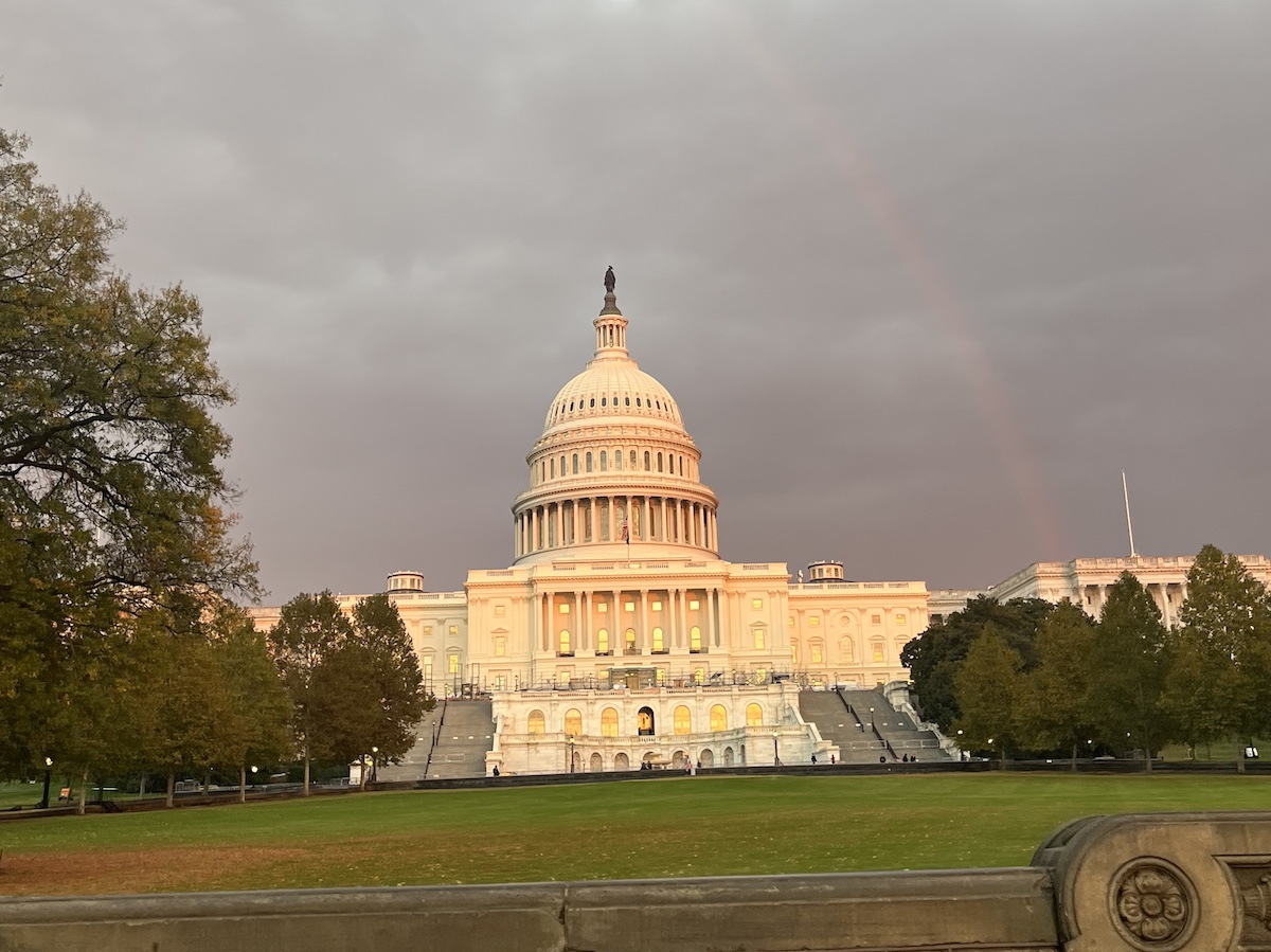 The U.S. Capitol building at sunset, under a cloudy sky with a partially visible rainbow in the background.