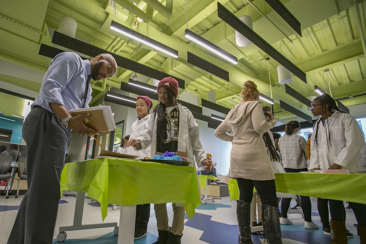 A man holding a folder speaks to students wearing lab coats and protective gloves in a modern classroom or lab setting. Other students are engaged in various activities around tables.