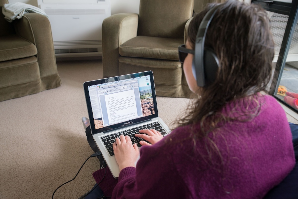 Person with long hair in a purple sweater and headphones works on a laptop in a living room setting. Two armchairs and a window are visible in the background.