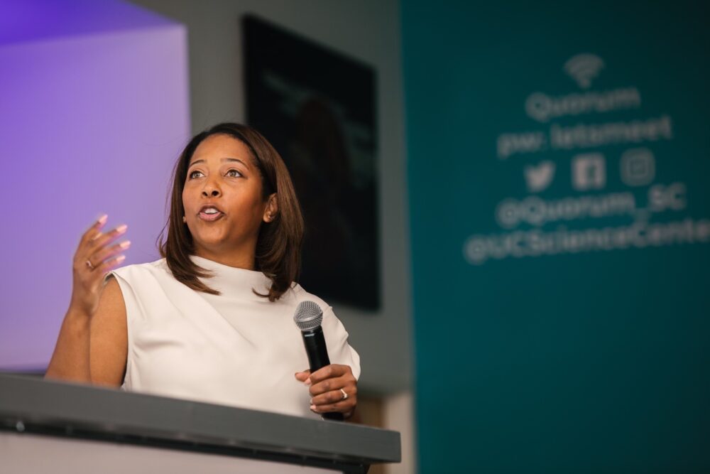 A woman in a white dress speaks into a microphone during a presentation. A teal-colored banner with social media handles is partially visible in the background.