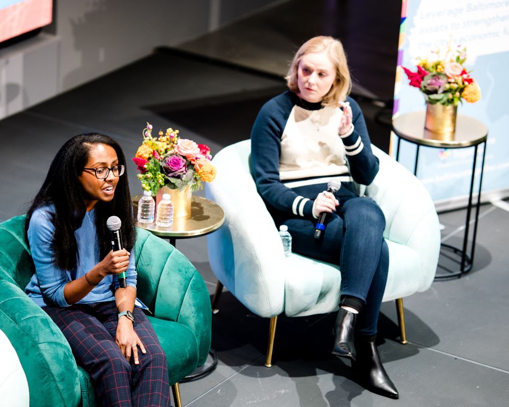 Two women seated in armchairs on a stage. One woman is speaking into a microphone, the other is holding a microphone and looking at her. A table with flowers is between them.