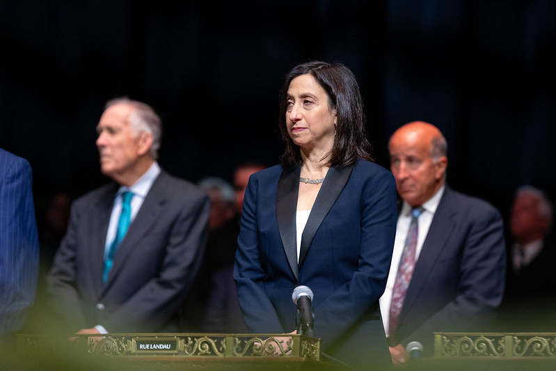 A woman in a blue suit stands solemnly in front of a microphone.
