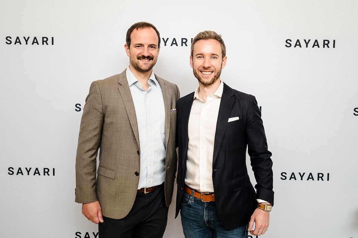 Two men wearing suit jackets and standing in front of a white backdrop.
