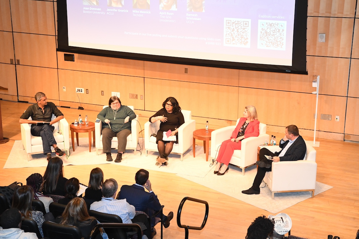 Five panelists on white chairs on brown wood stage