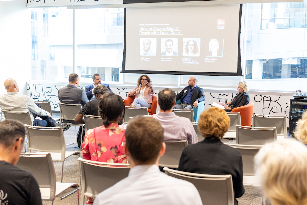 A group of people sitting in a panel discussion session. The sign above the presenters says "How to create global reach with local gains."