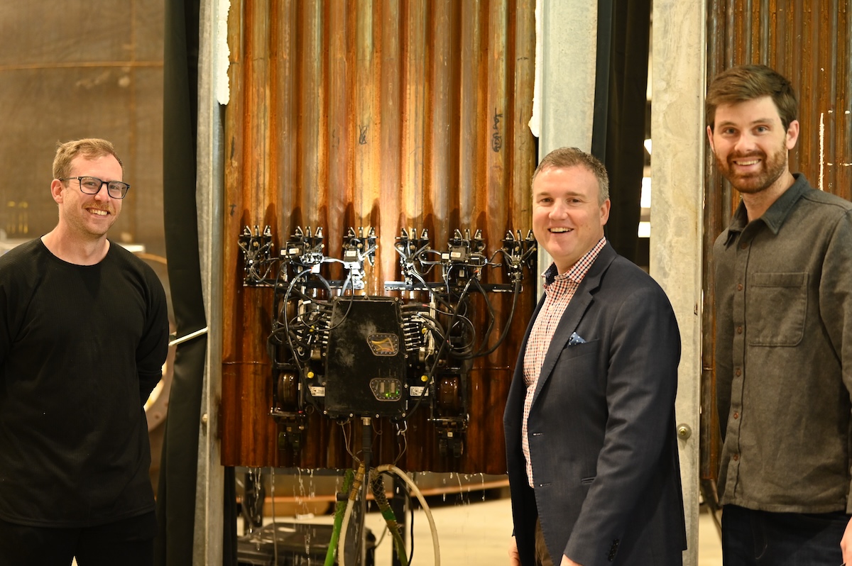 Three men standing in front of a machine, smiling.