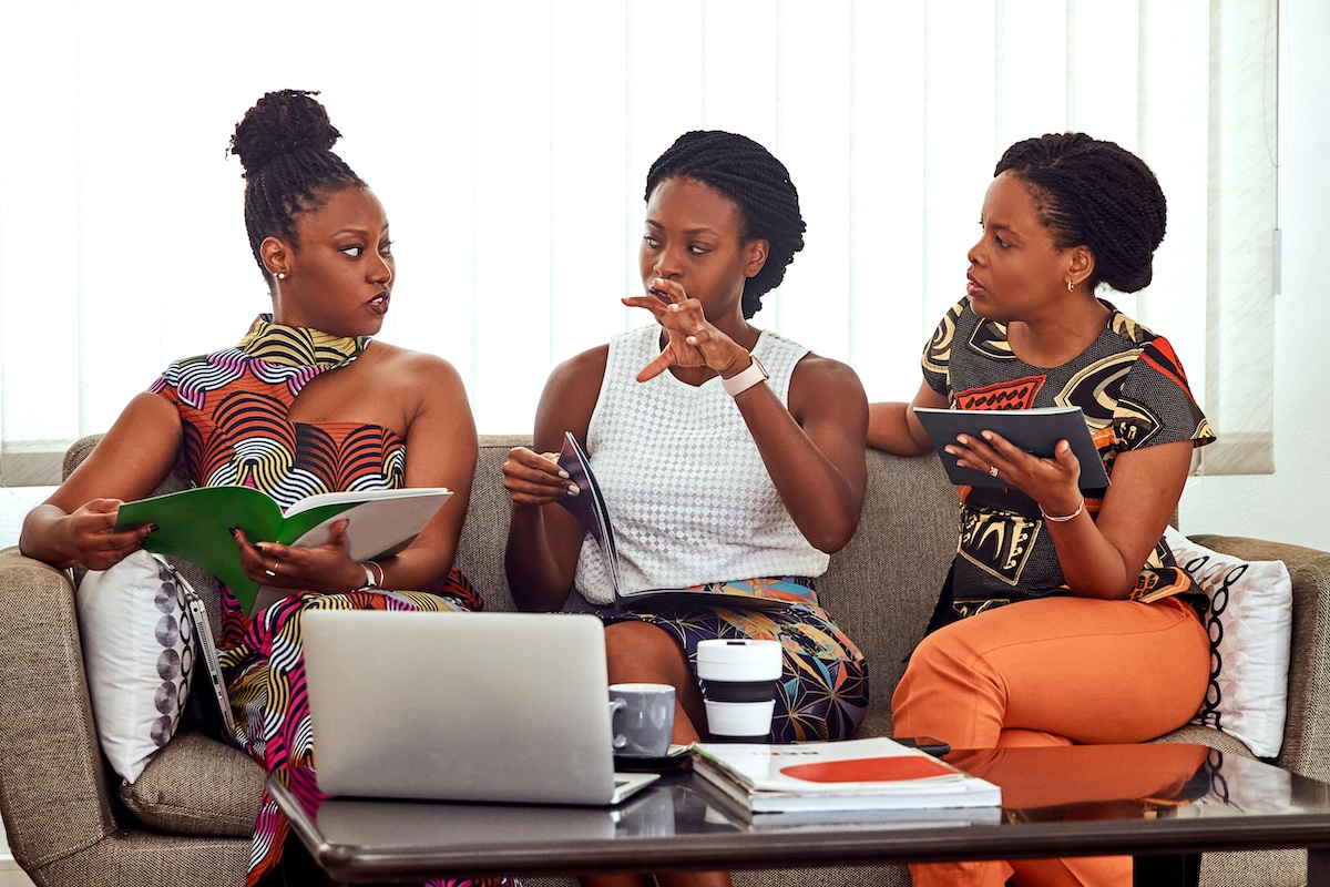 Three Black women sitting on a couch with laptop and booklets.