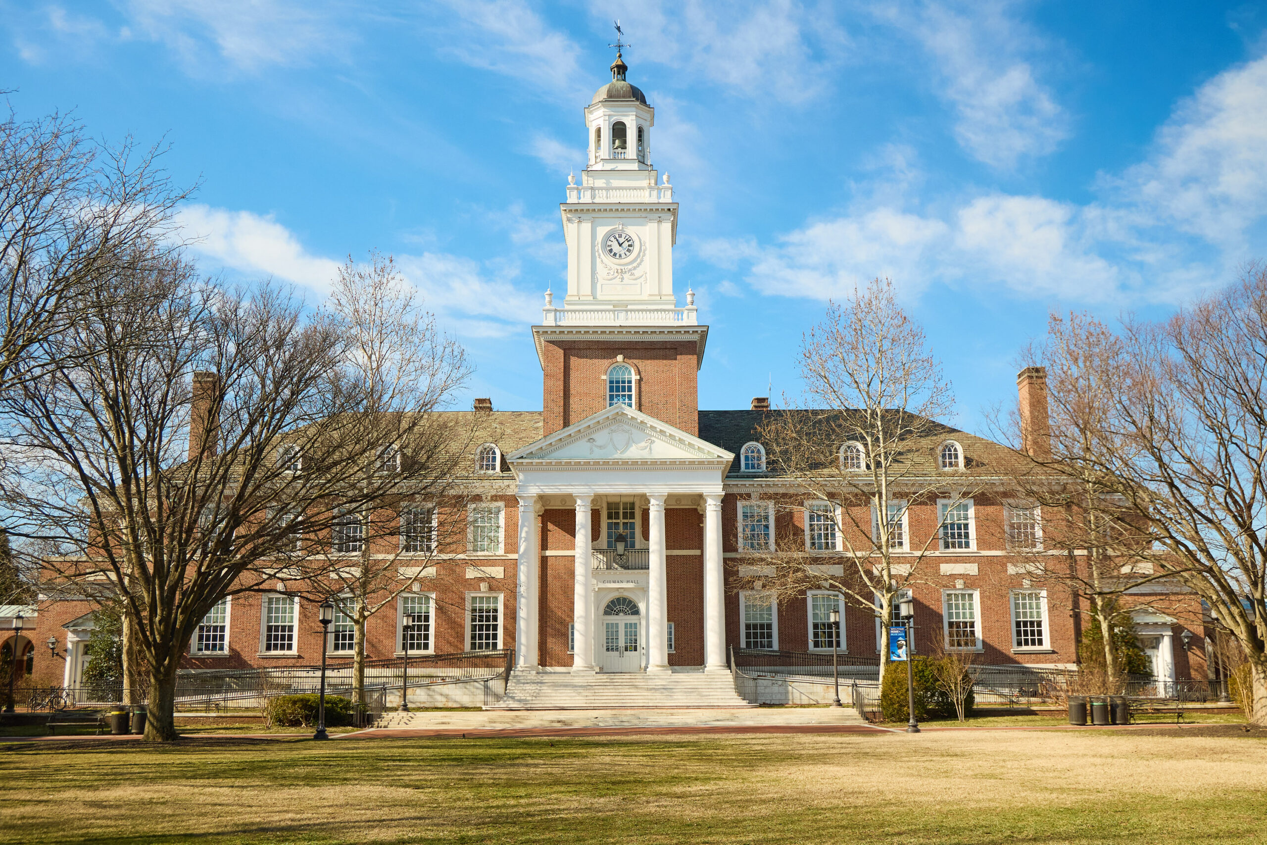 A large brick academic building with a central clock tower, white columns, and a pediment. The building is surrounded by leafless trees and a grassy courtyard under a blue sky.