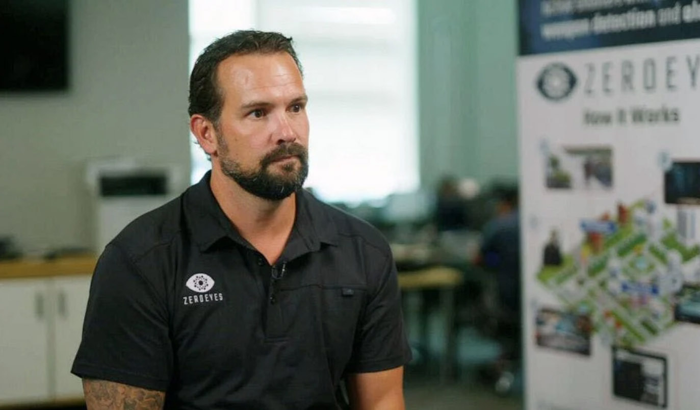 A man with a beard sitting in an office environment, wearing a black shirt with a logo, with promotional material in the background.
