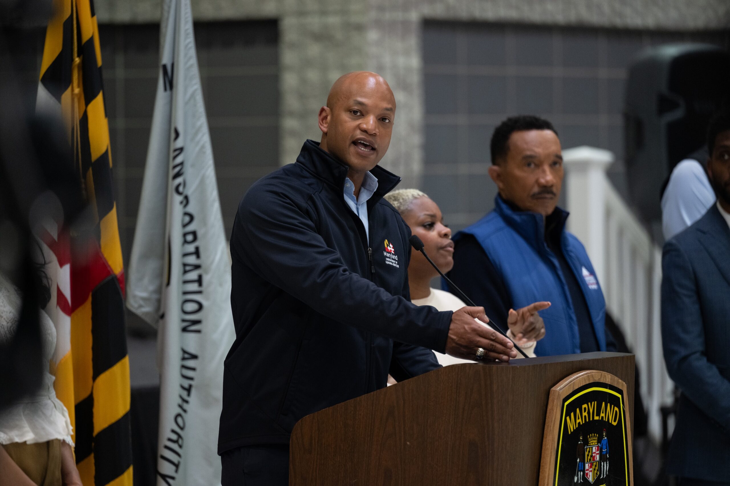 Gov. Wes Moore speaks in navy-black jacket behind brown podium