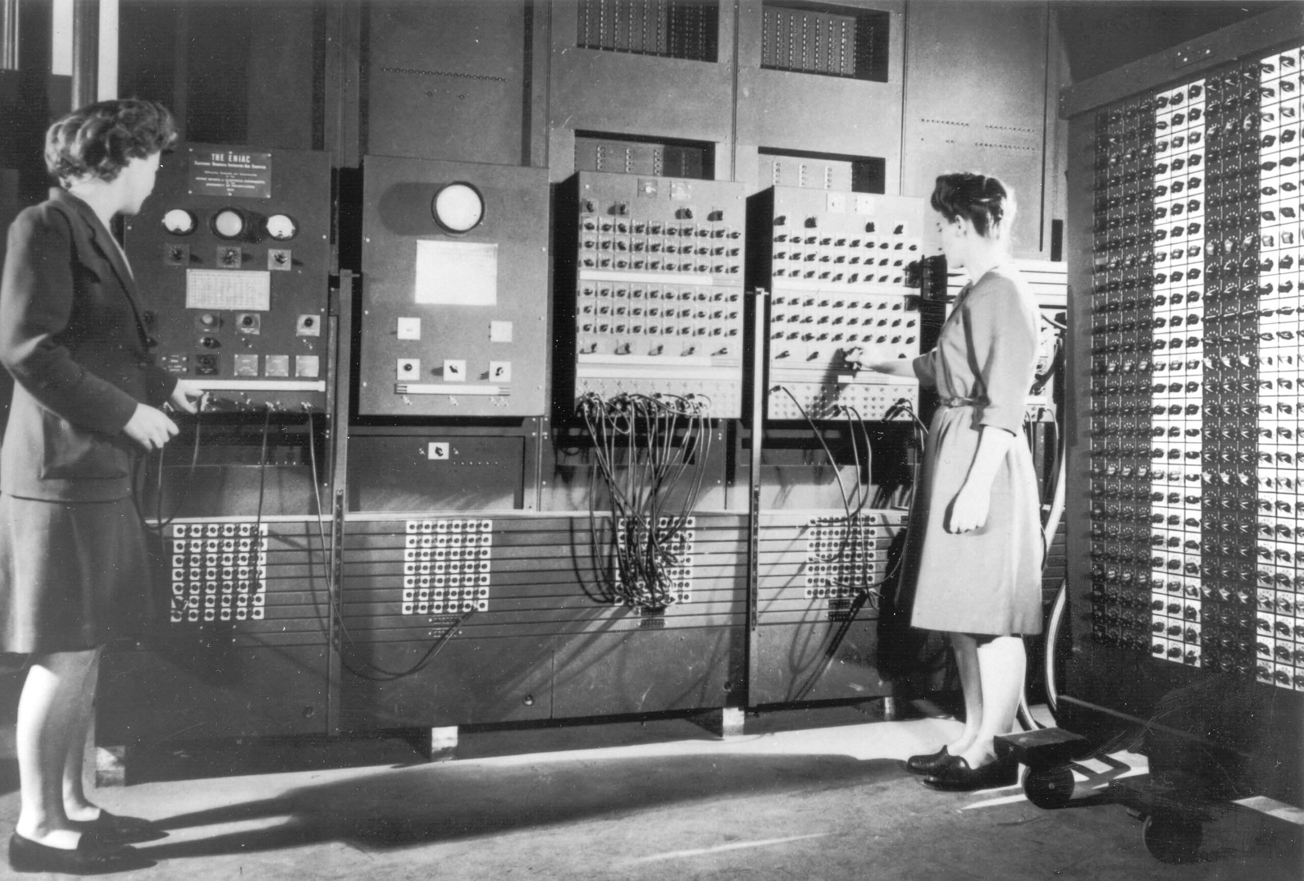 Two women operating an early computer with a bank of plugboards and switches.