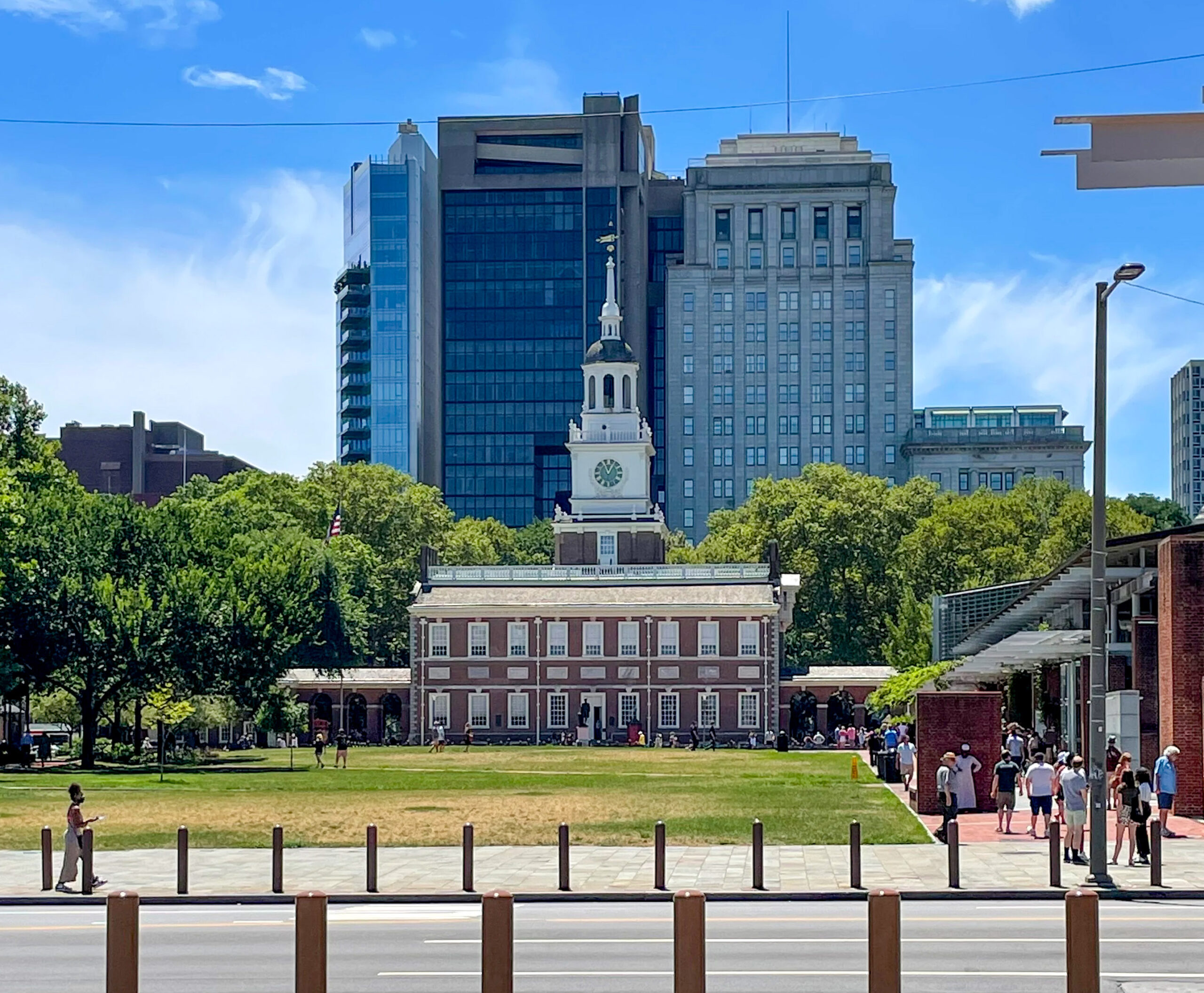 Historic building with clock tower in the foreground, modern skyscrapers in the background, and tourists milling about on a sunny day.