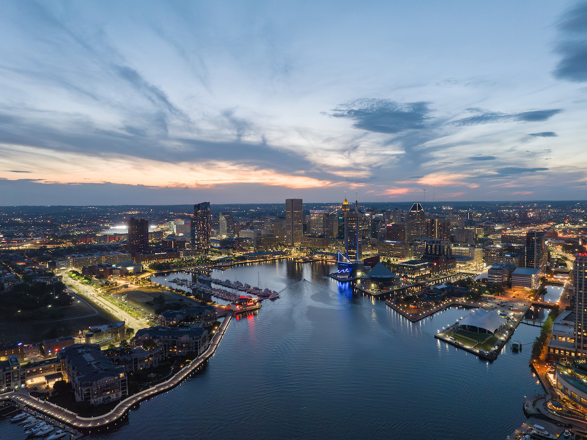 Aerial view of a city skyline at dusk, featuring illuminated buildings, waterfront, and a harbor with boats under a partly cloudy sky.