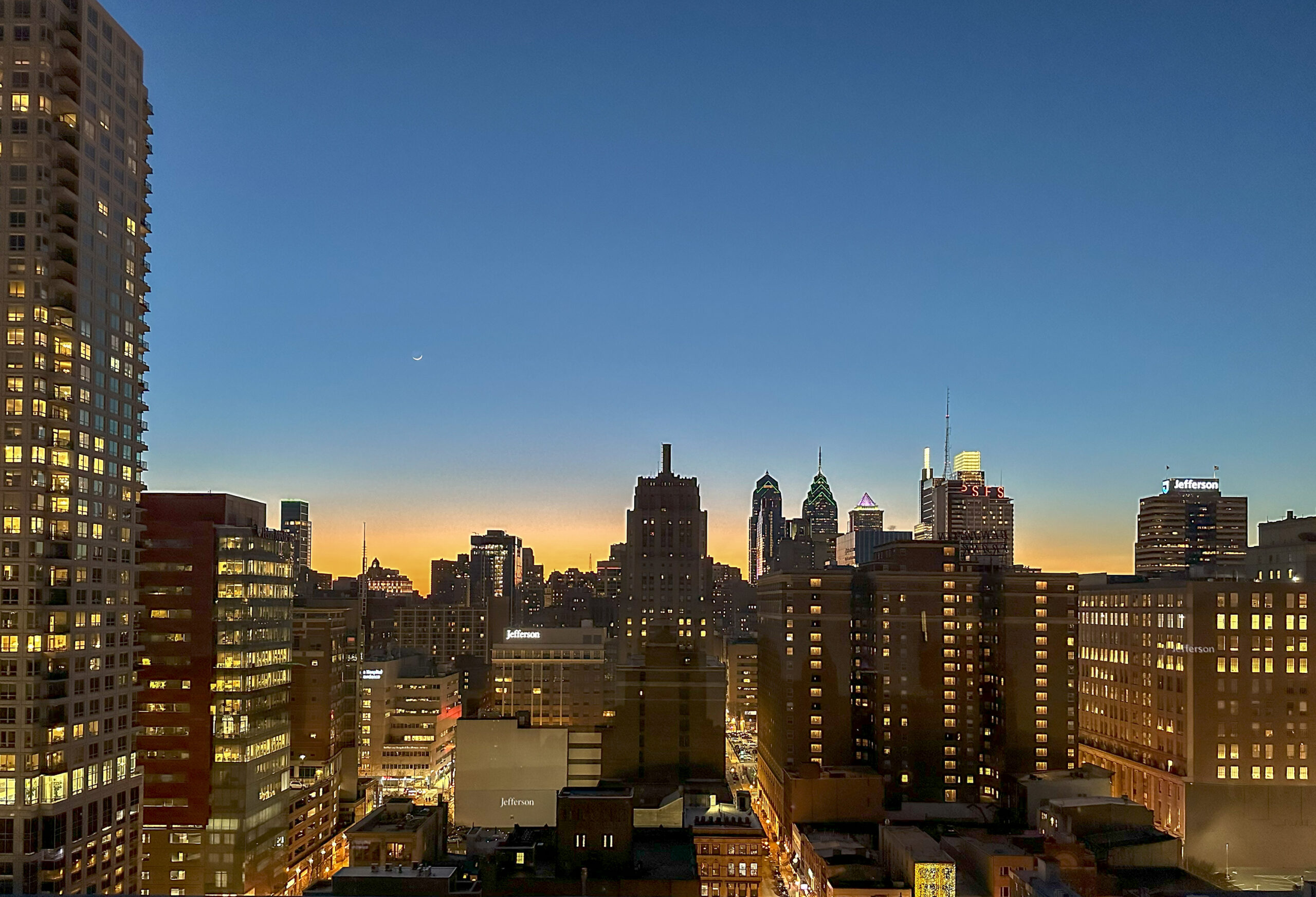 Cityscape at dusk with illuminated buildings and a clear sky, featuring a crescent moon above a silhouette of skyscrapers.