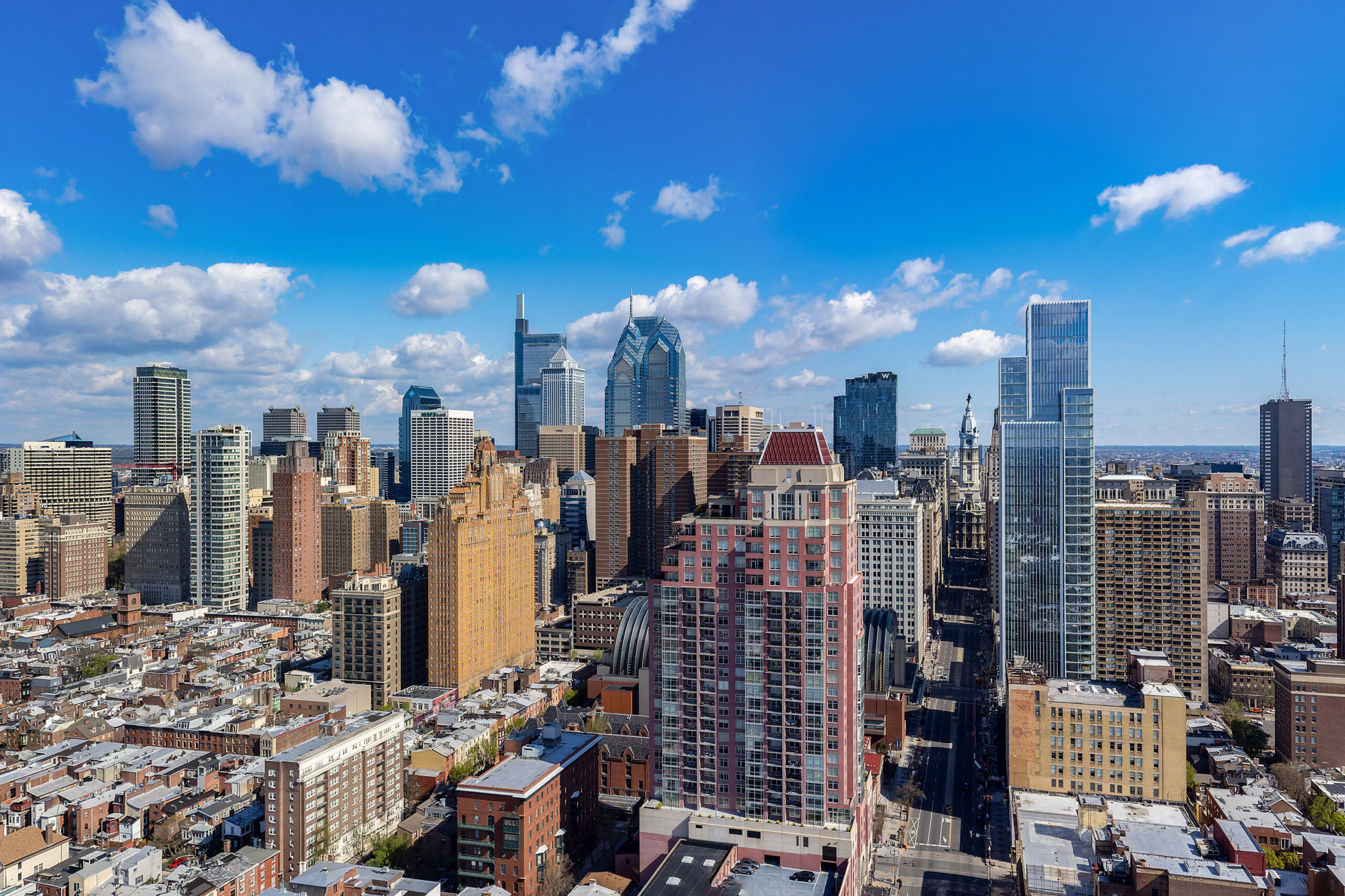 Aerial view of Center City Philadelphia on a clear day, showcasing skyscrapers and dense urban architecture under a blue sky with scattered clouds.