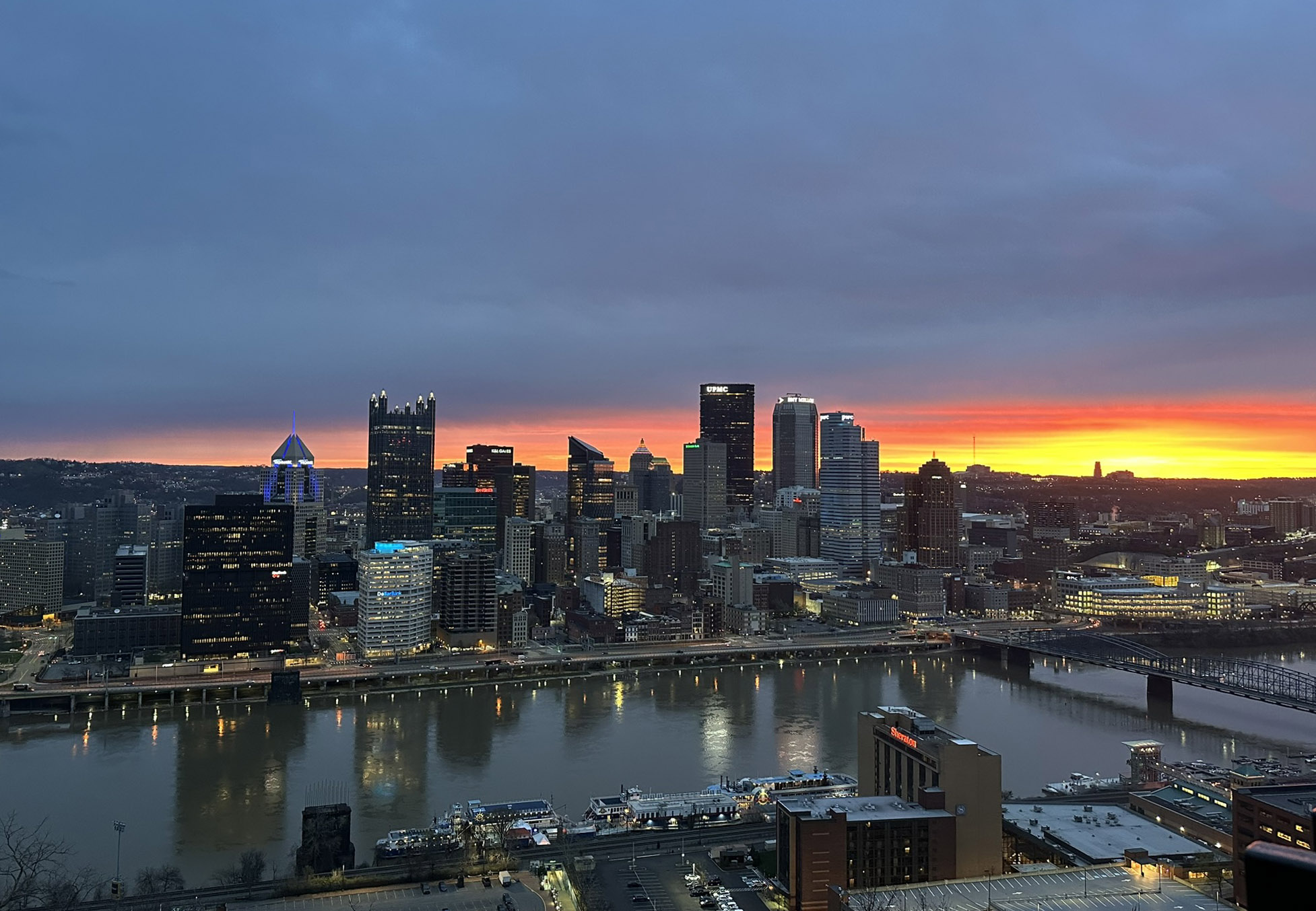 A city skyline at dusk with a mix of modern high-rise buildings reflecting on a river. The sky displays a vibrant sunset with hues of orange and yellow near the horizon.
