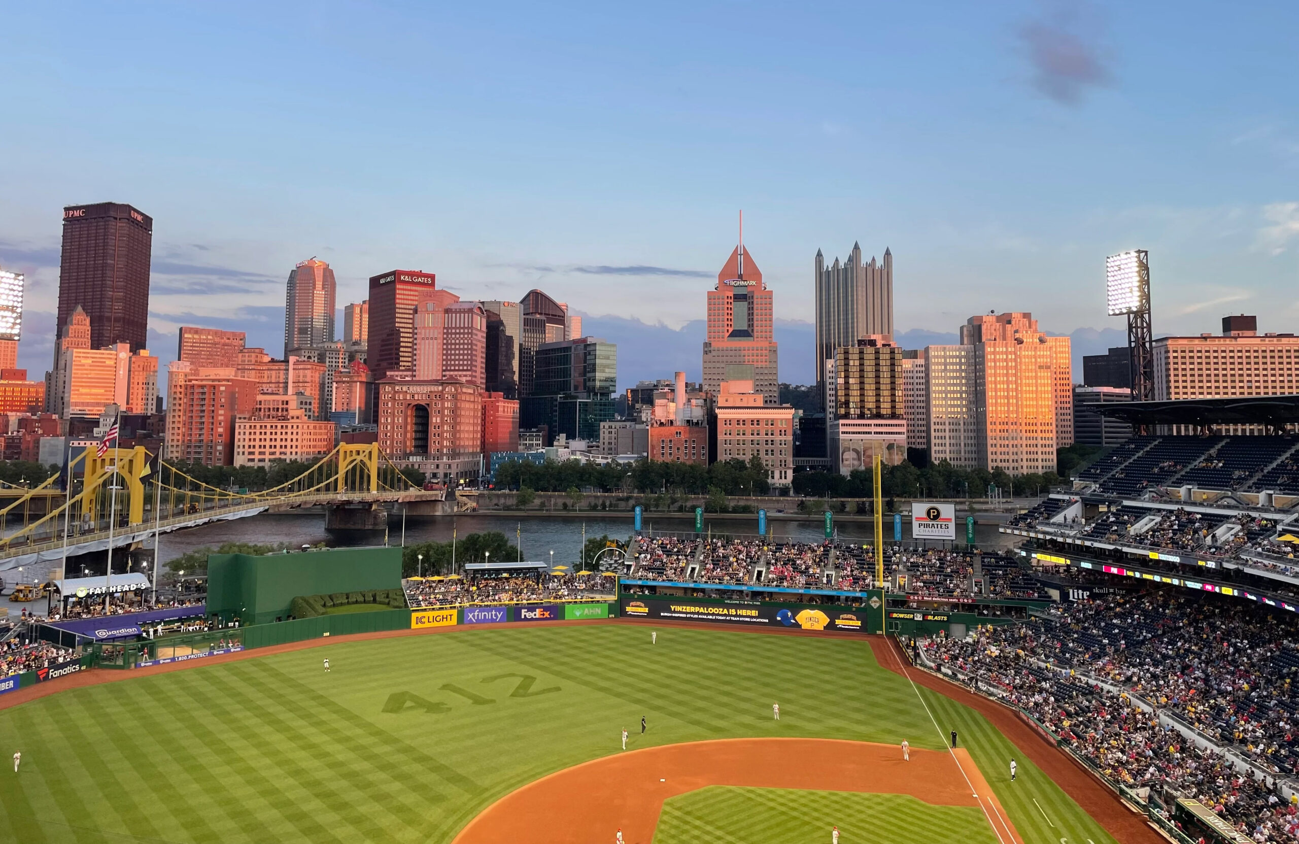 View of a baseball game in progress at a stadium with downtown Pittsburgh skyline in the backdrop during sunset.