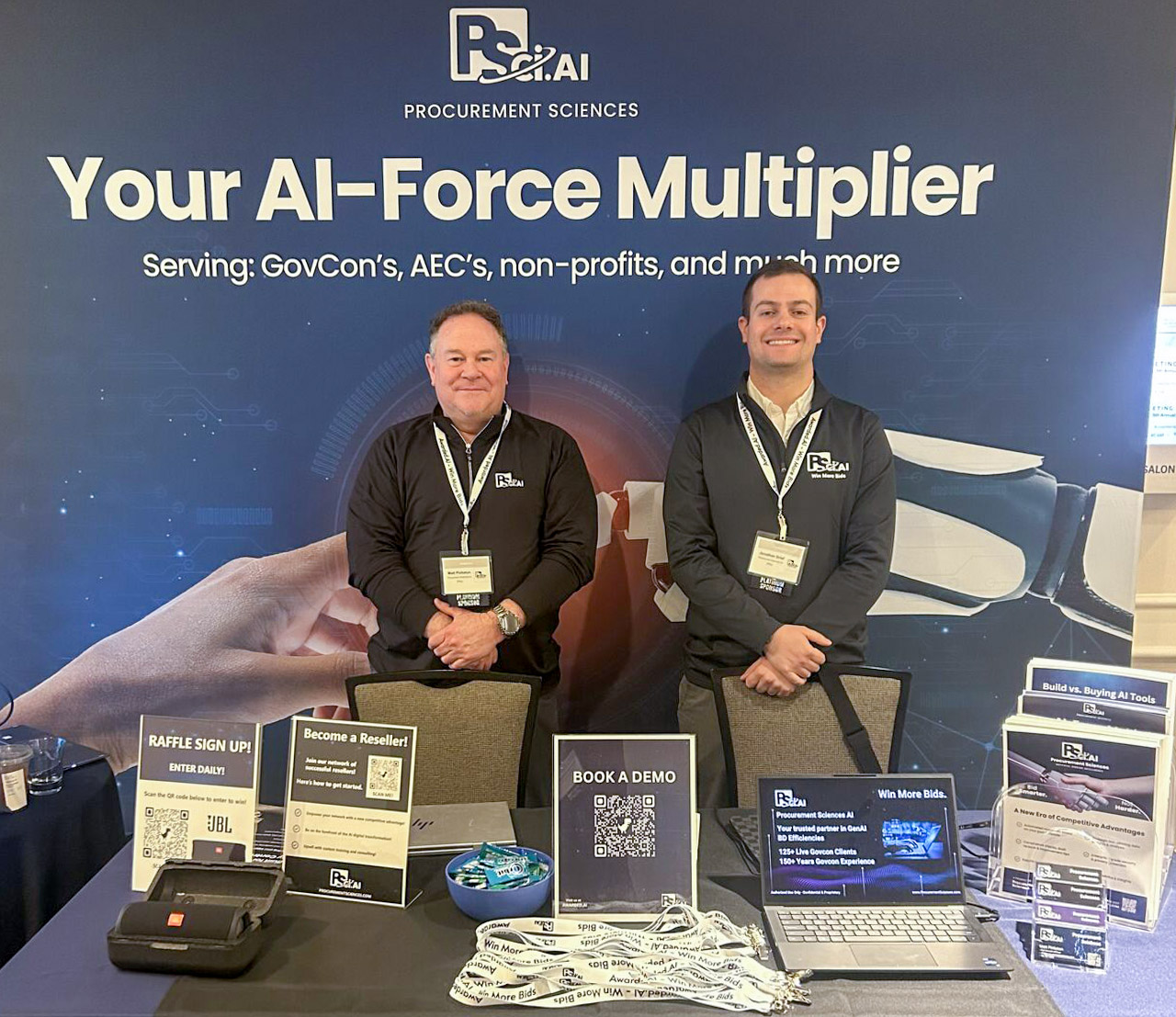 Two men seated at a promotional booth for ps.ai with brochures, a raffle sign-up, and a demo booking area.