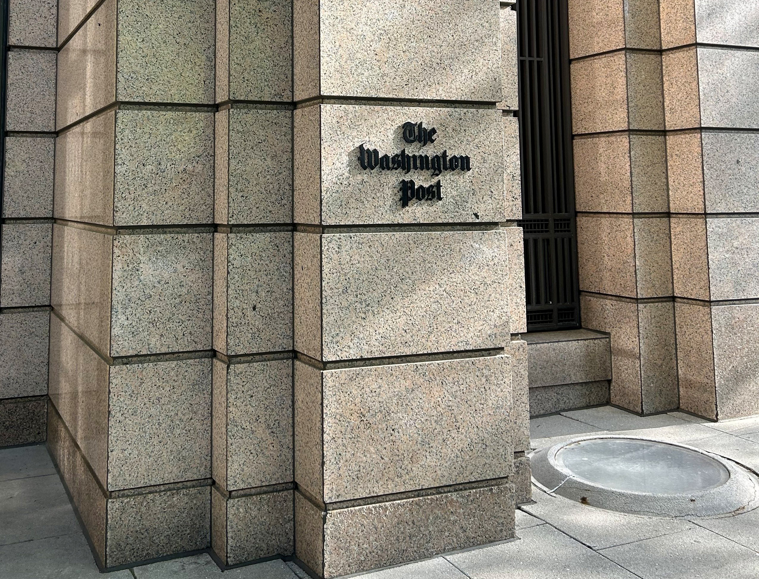 The entrance corner of the Washington Post building, featuring the paper's name on the wall above a dark doorway, framed by granite tiles.