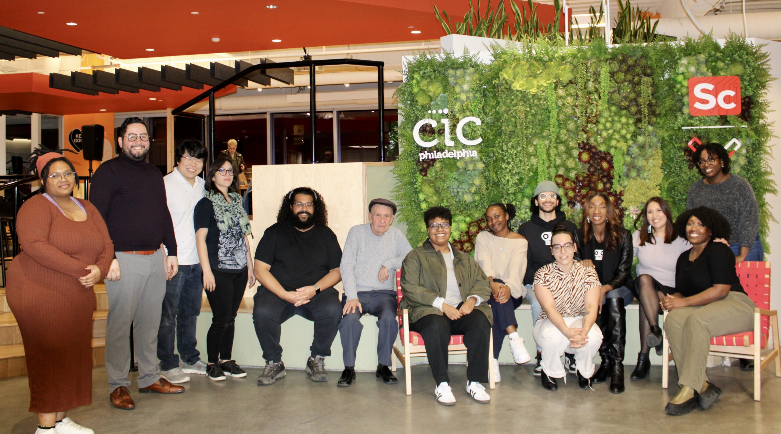 Group of 14 people posing for a photo in front of a green foliage wall with CIC Philadelphia and Science Center logos. Some are standing while others are seated. Indoor setting with stairs in background.