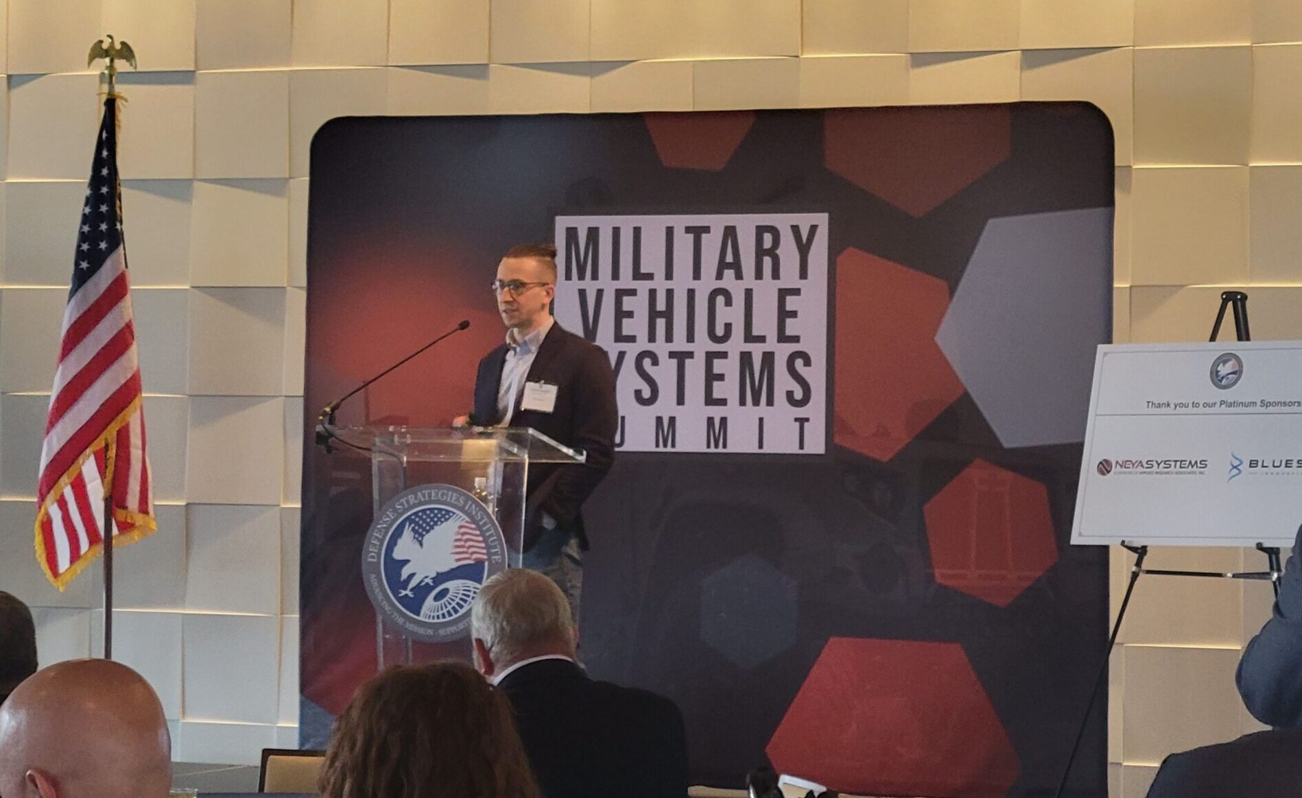 A man speaks at a podium in front of a sign that reads "Military Vehicle Systems Summit." An American flag is on the left, and audience members are seated in the foreground.