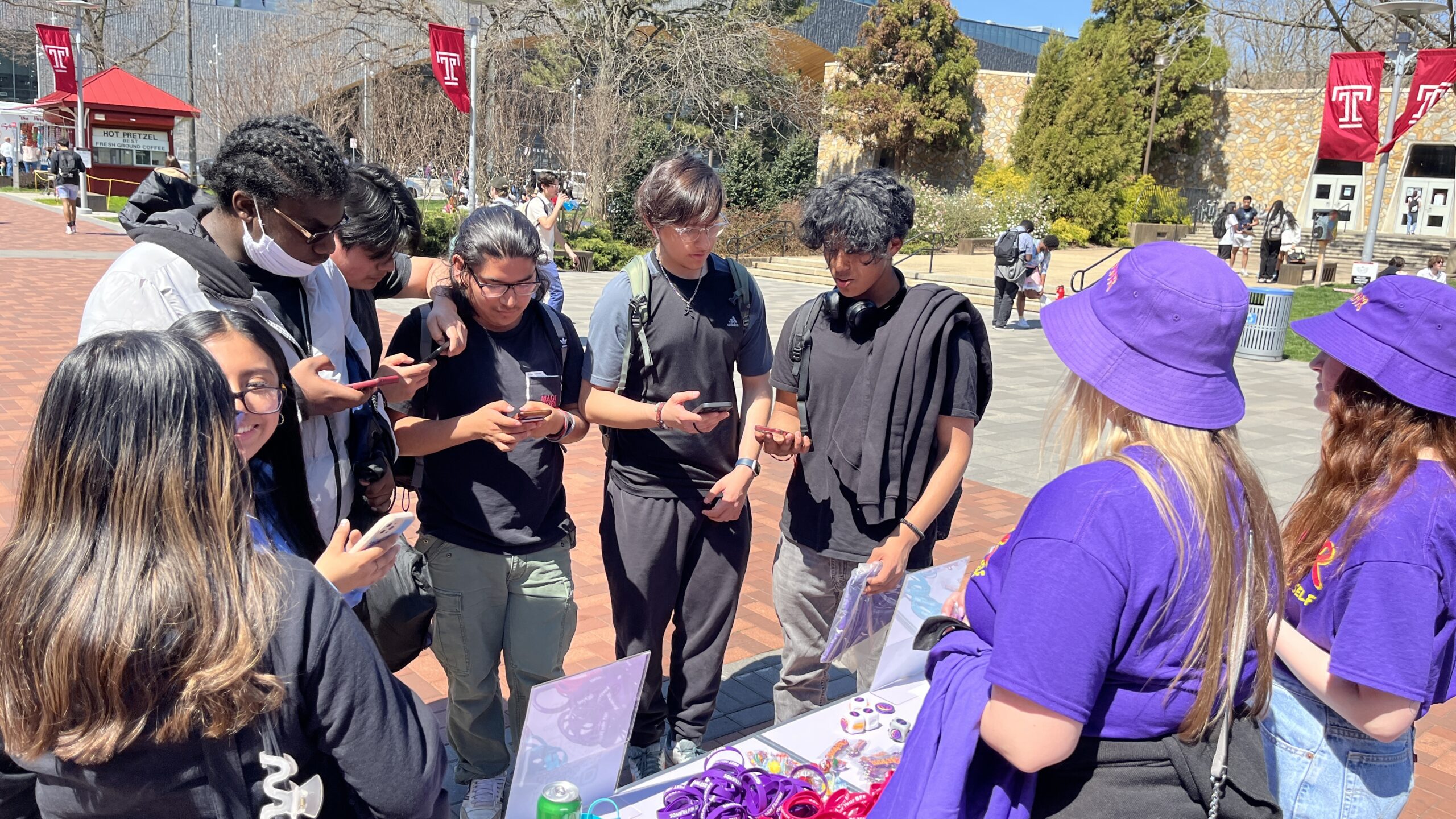A group of students stands around a table outside on a sunny day, looking at their phones and at items on the table. Two individuals in purple T-shirts and hats appear to be assisting them.