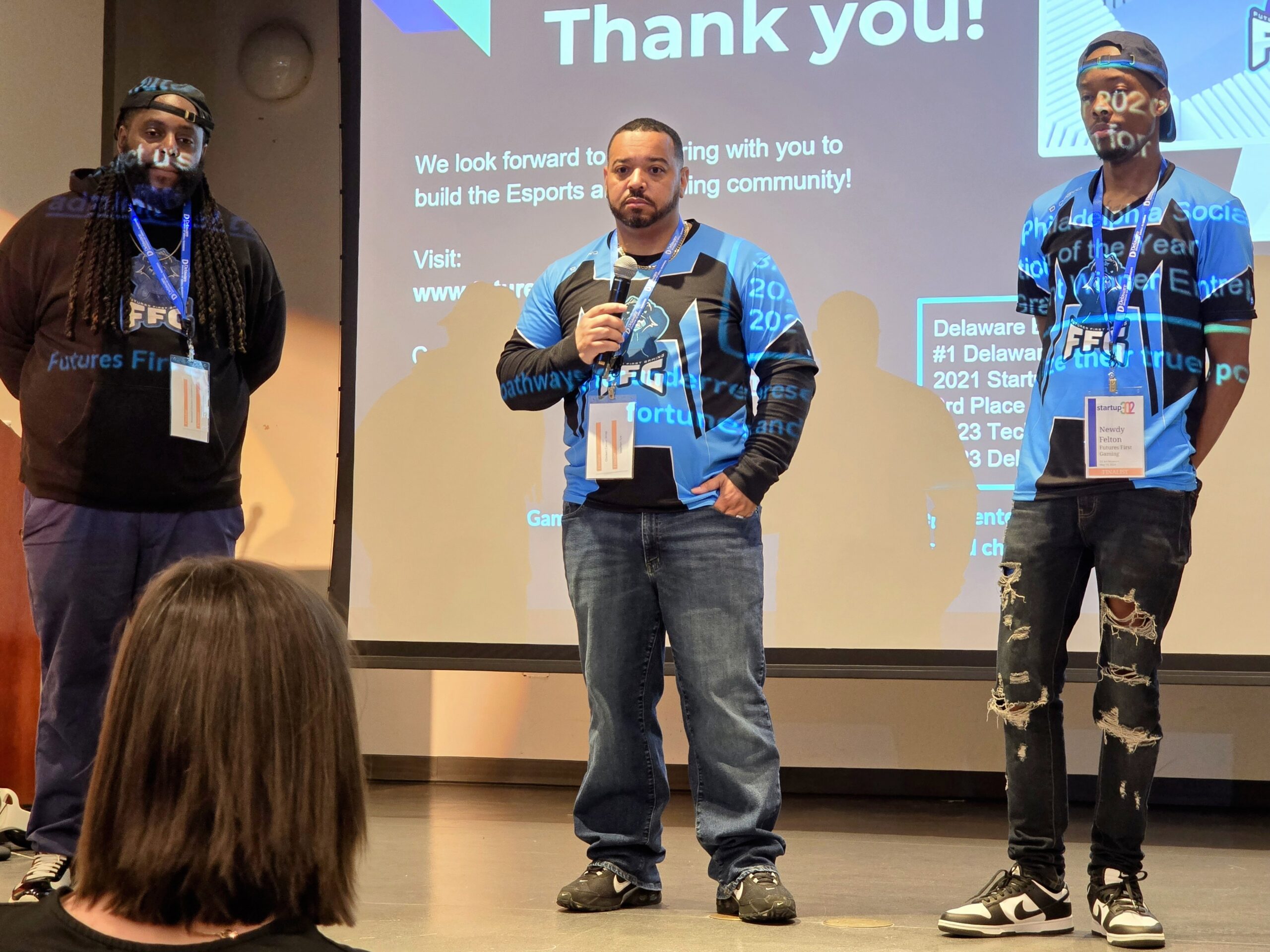 Three individuals stand on stage, with one speaking into a microphone. They are in front of a projected slide that reads "Thank you!" and are wearing blue-themed casual attire.
