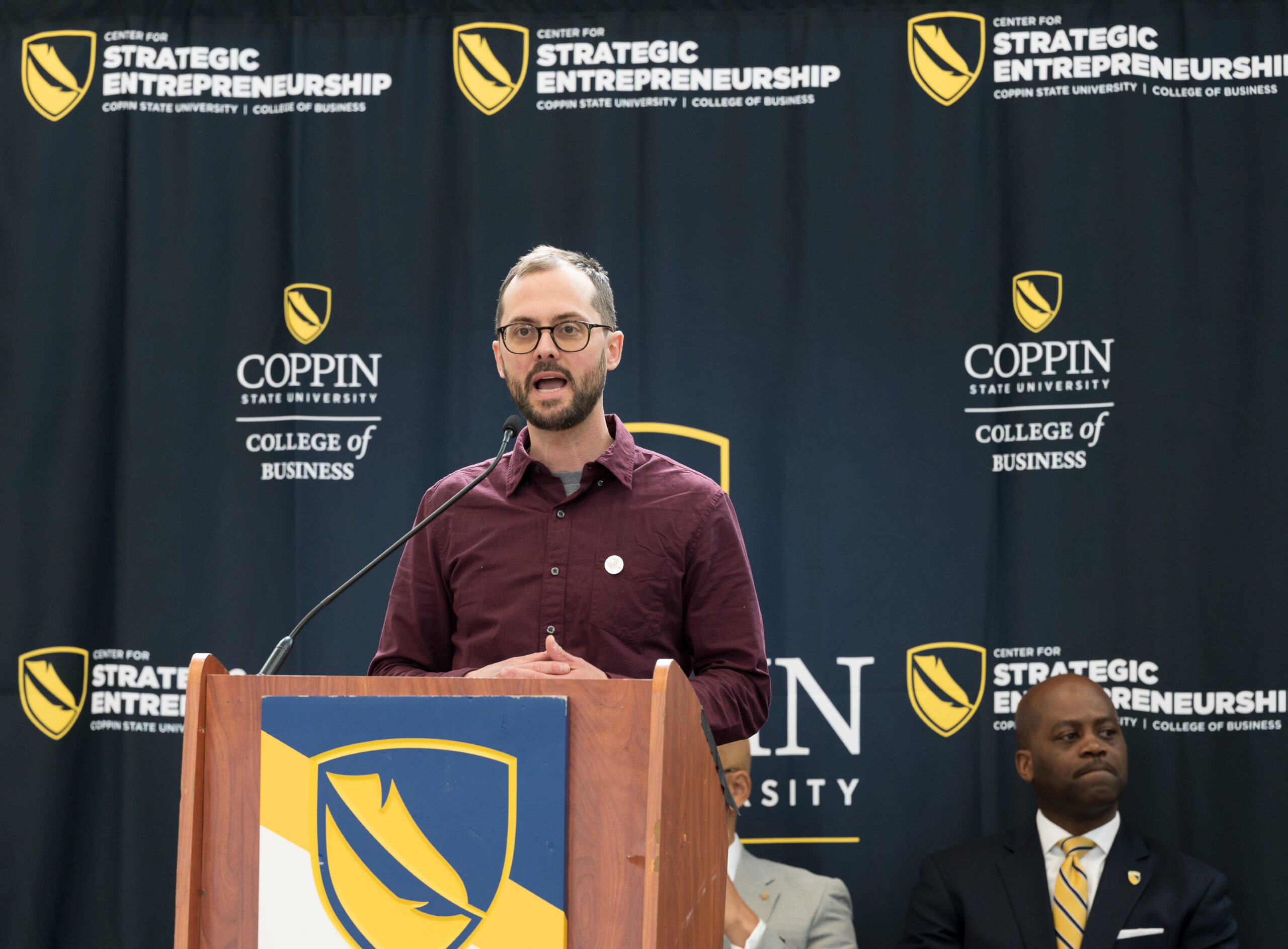 White man in brown shirt stands behind brown podium.