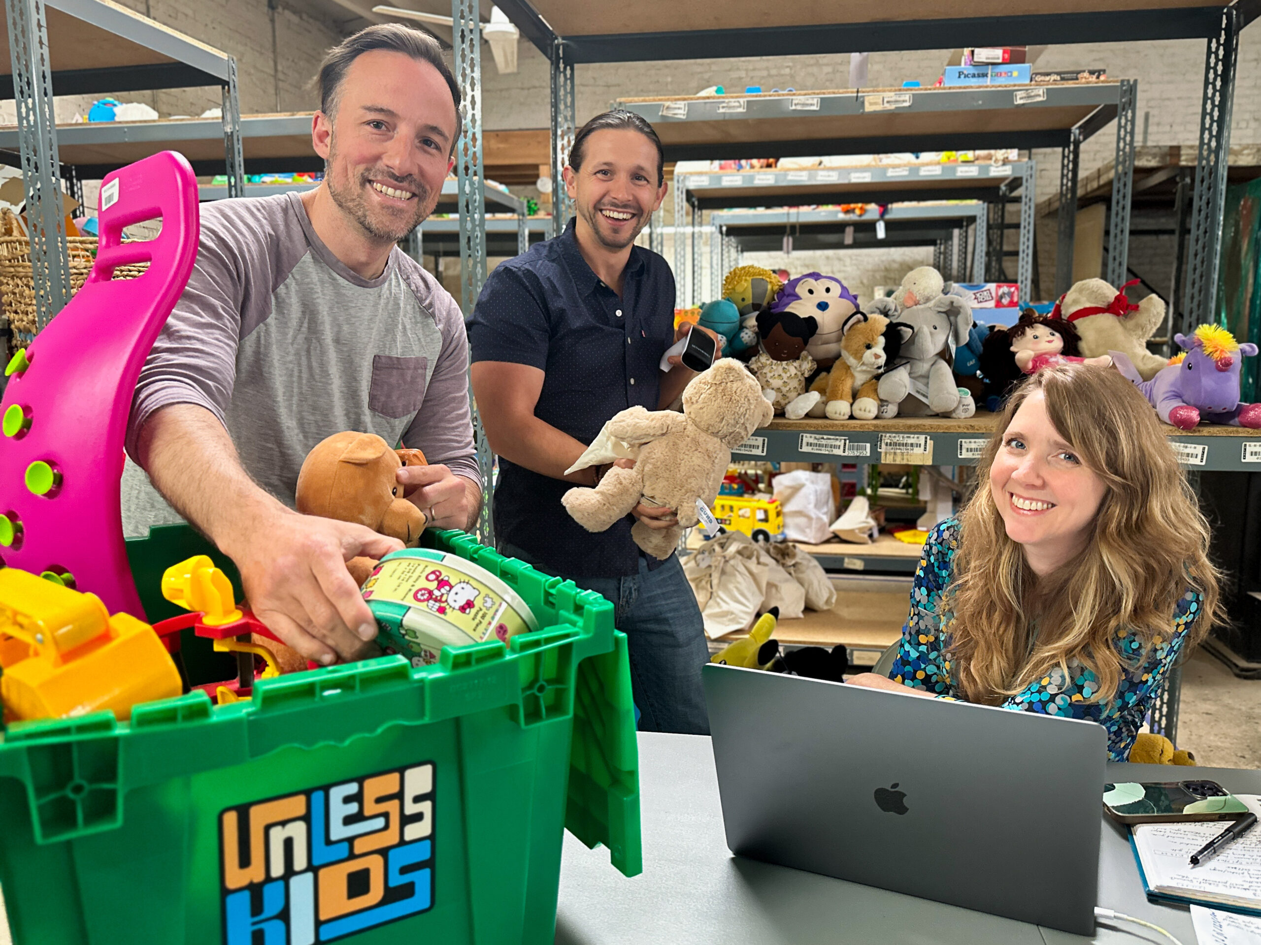 Three people are organizing toys in a storage area. A man on the left is placing items in a green bin, another man in the center is holding a teddy bear, and a woman on the right is working on a laptop.