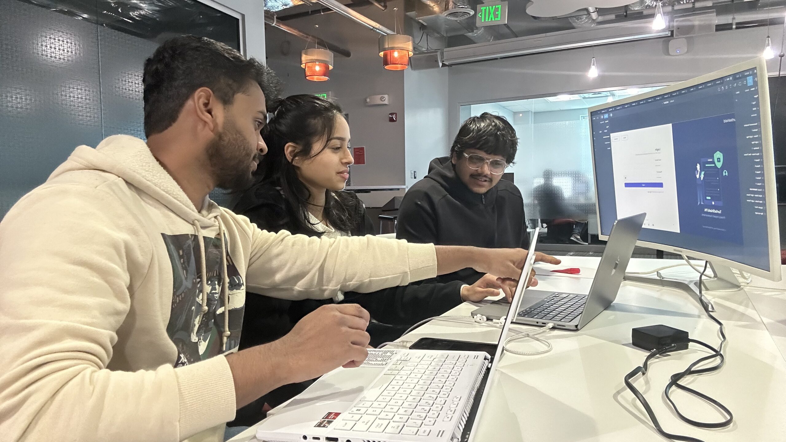 Three people, seated at a table with laptops, work together on a computer project. One of them points at the large monitor in front of them. An office setting with modern decor is visible in the background.