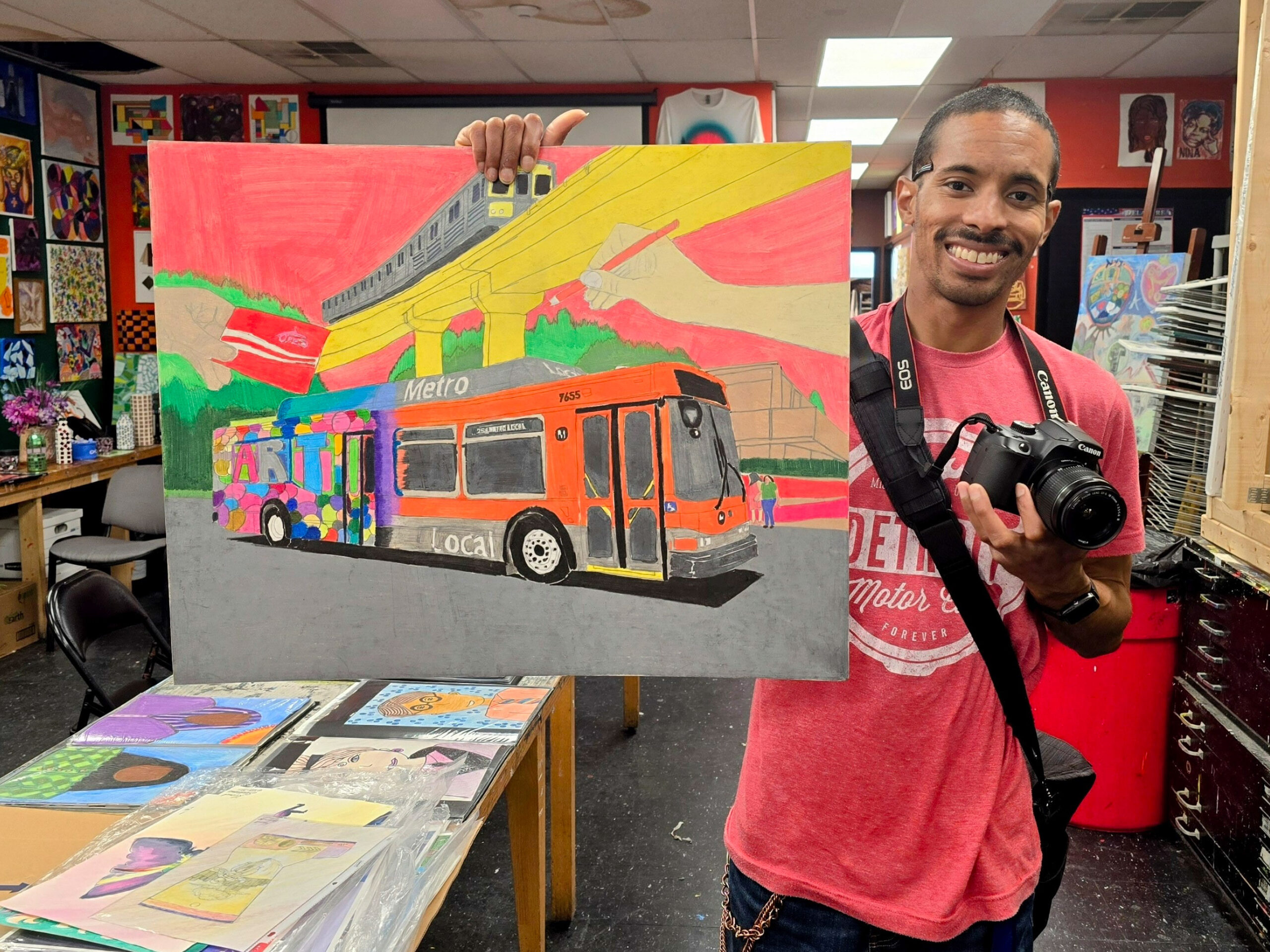 A man wearing a pink shirt holds a colorful artwork of a bus. He stands in an art classroom, smiling, with a camera around his neck.
