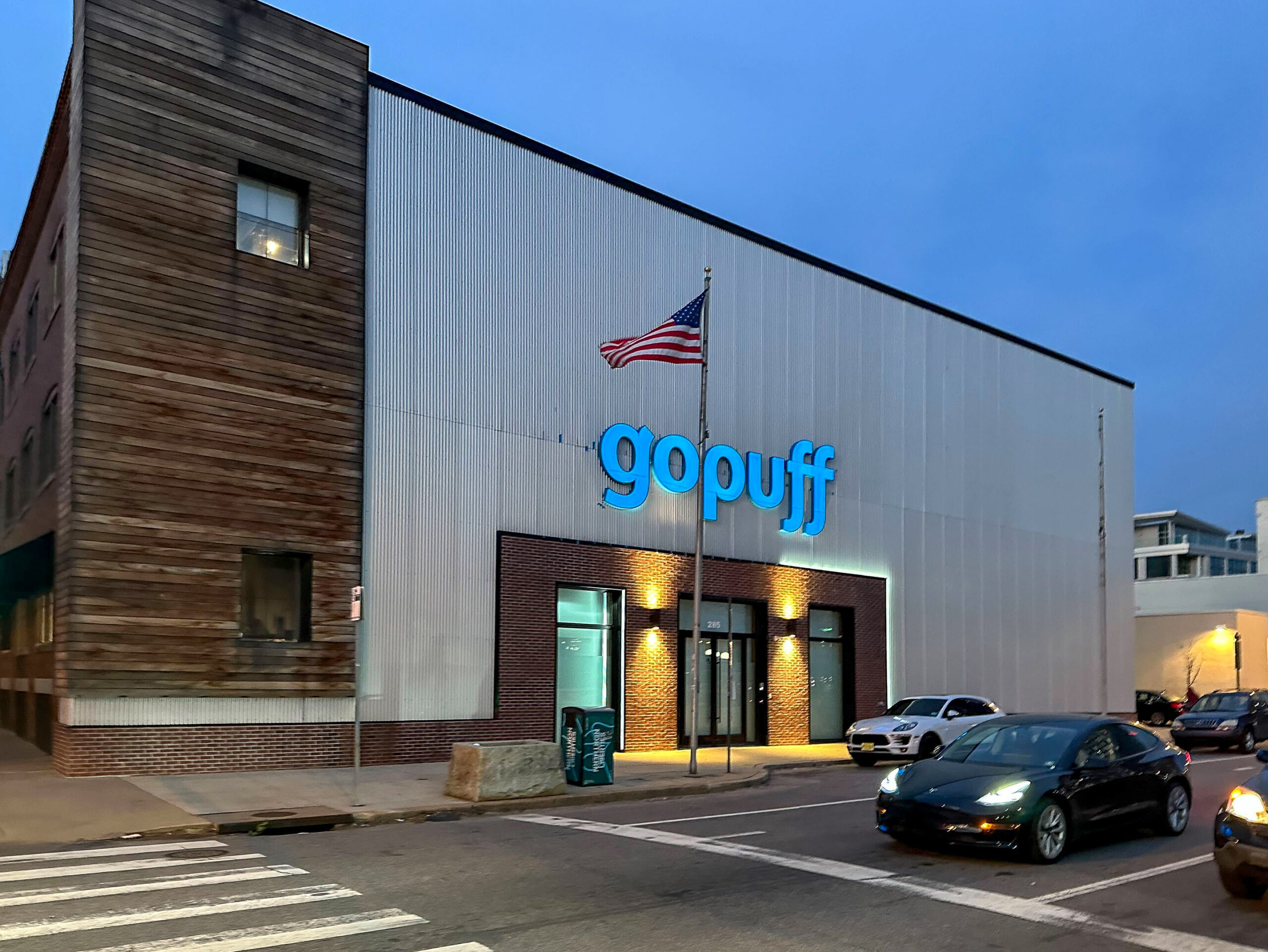 Street view of a Gopuff delivery service warehouse with an American flag on top, illuminated in the evening with cars parked nearby.